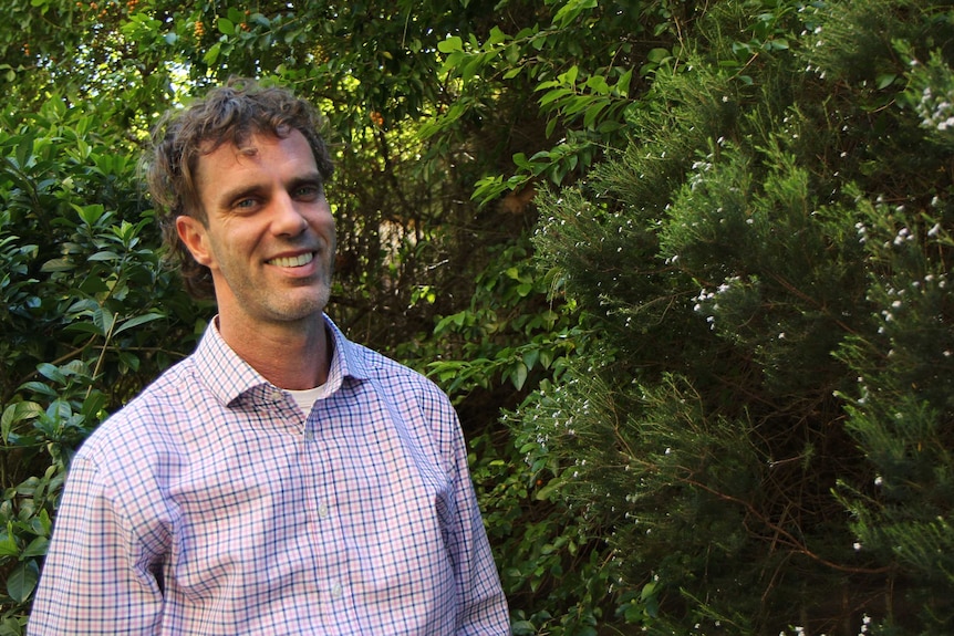 A man in a checked shirt stands in front of greenery.