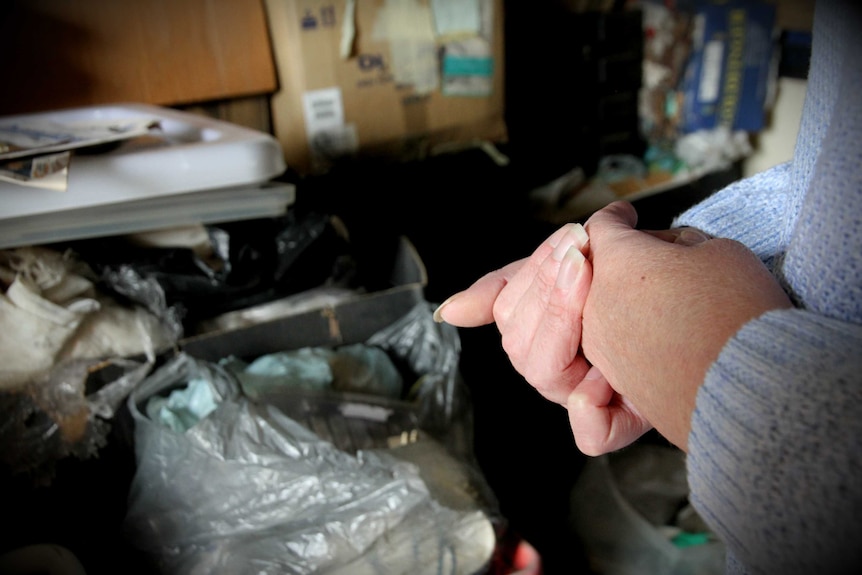 Close up of a woman's hands, clasped in her garage. Bags of garbage are in the background.
