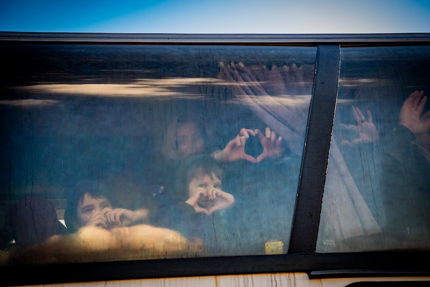 Women and children seen from outside a bus use their hands to make heart signs through the window