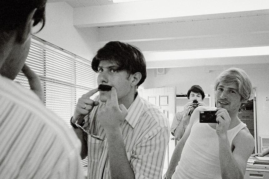In front of a mirror two men in short sleeve shirts pose with fake moustaches, between them a man holds up small film camera.