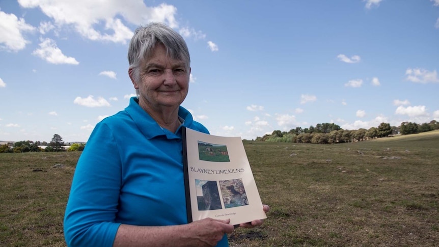 An older woman standing in a paddock holding a book called Blayney Limekilns