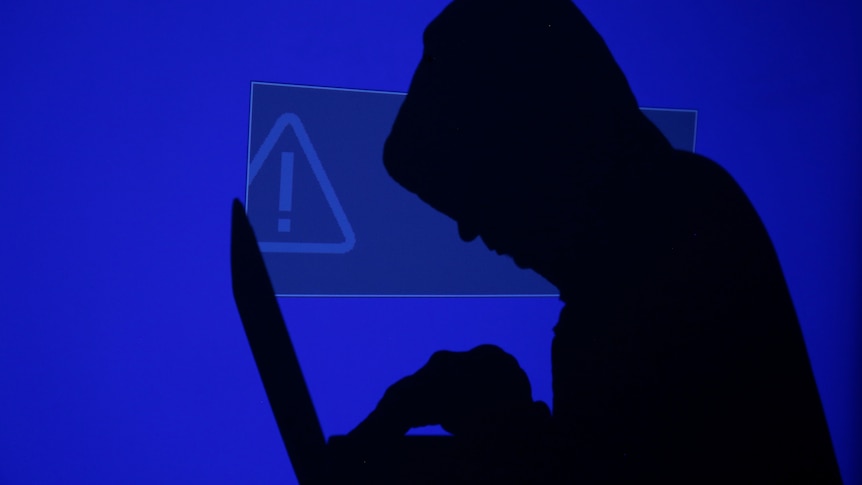 A hooded man holds a laptop computer as blue screen with an exclamation mark is projected on him