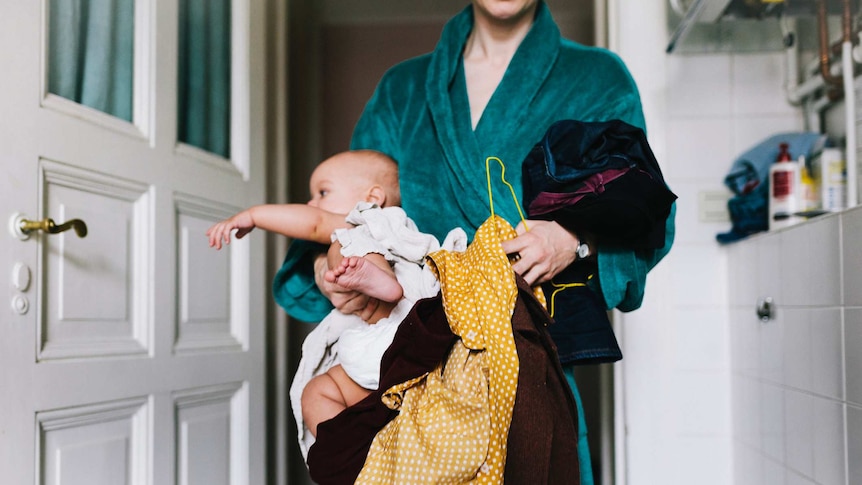 A woman in a robe holds bundles of clothes and her baby