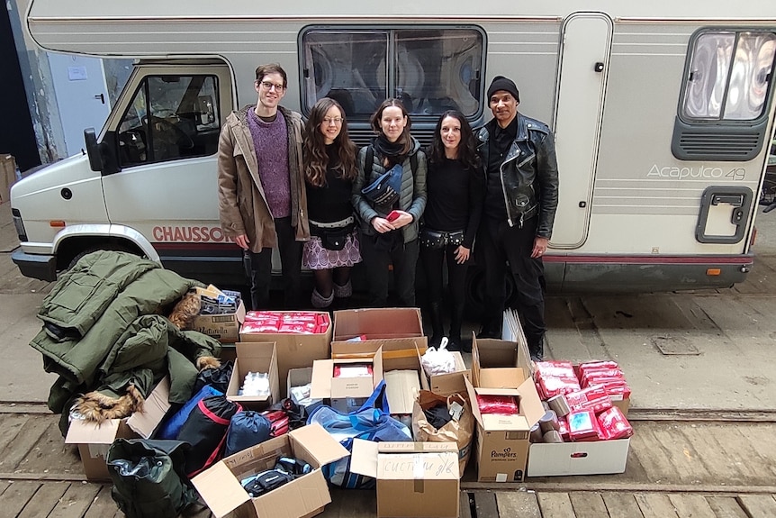 A group of young people standing in front of a campervan with boxes and coats in front of them. 