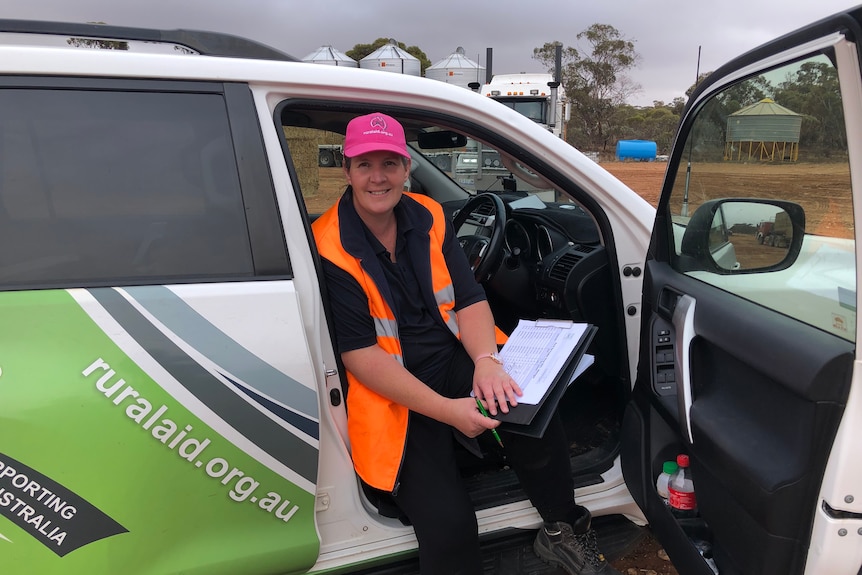 A woman wearing a hi-vis orange vest and pink cap sits in driver's seat of her car, holding a clipboard