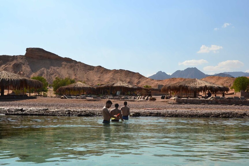 Three male tourists in the water, with huts and hills in the background
