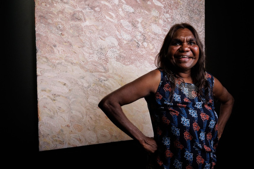 A beaming woman standing in front of a large artwork hung on a black wall with her hands on her hips.