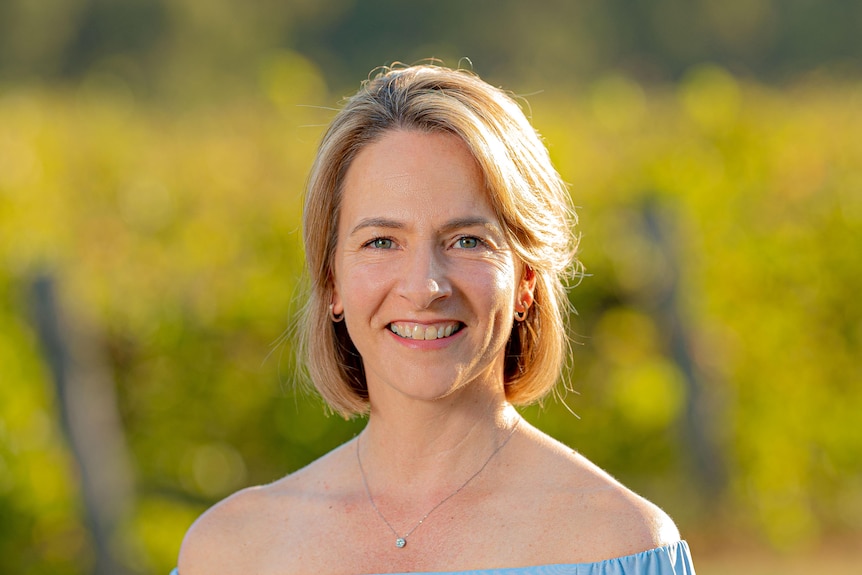 A blonde woman is smiling at the camera among vines.