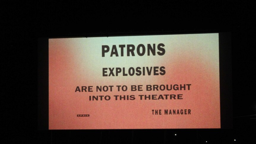 'Patrons explosives are not to be brought into this theatre' message sits against an orange background on a theatre screen