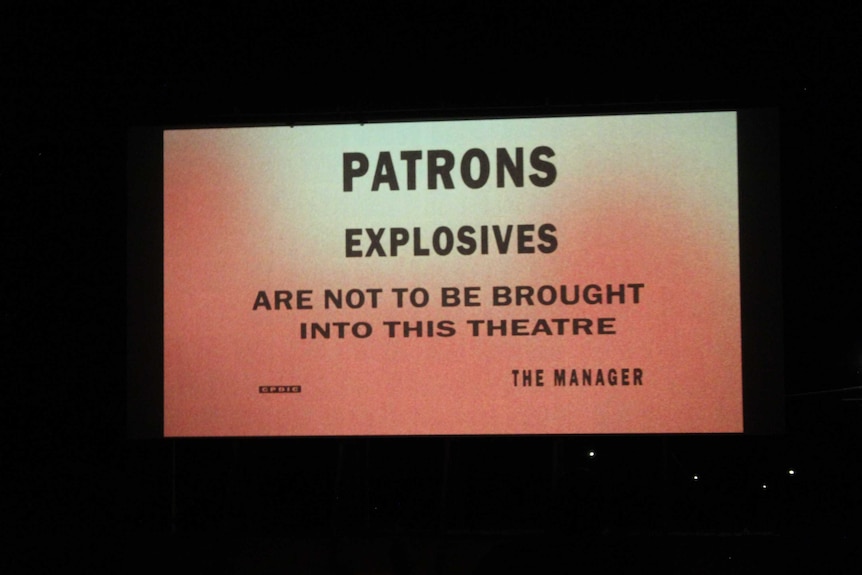 'Patrons explosives are not to be brought into this theatre' message sits against an orange background on a theatre screen