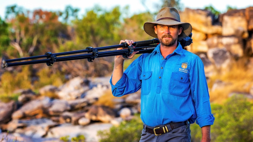 A man wearing an akubra stands amid a rocky outback landscape holding a camera and tripod over his shoulder.