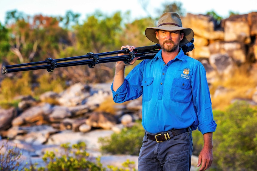 A man wearing an akubra stands amid a rocky outback landscape holding a camera and tripod over his shoulder.