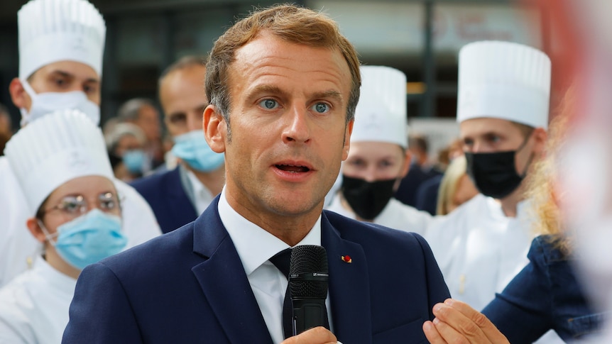 French President Emmanuel Macron wears a blue suit and holds a microphone.