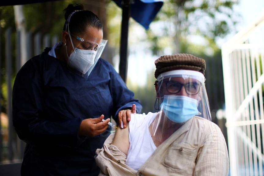 A healthcare worker in scrubs, face mask and shield inserts a needle into a man's arm. He is also wearing face mask and shield