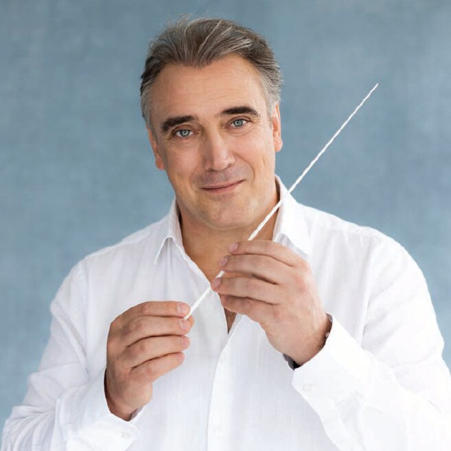 Jaime Martin, in a white collared shirt with two buttons undone, is holding a white conductor's baton in front of him