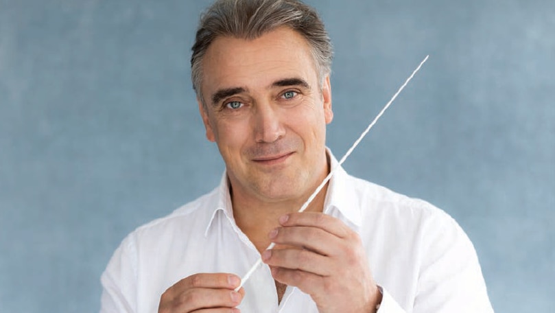 Jaime Martin, in a white collared shirt with two buttons undone, is holding a white conductor's baton in front of him