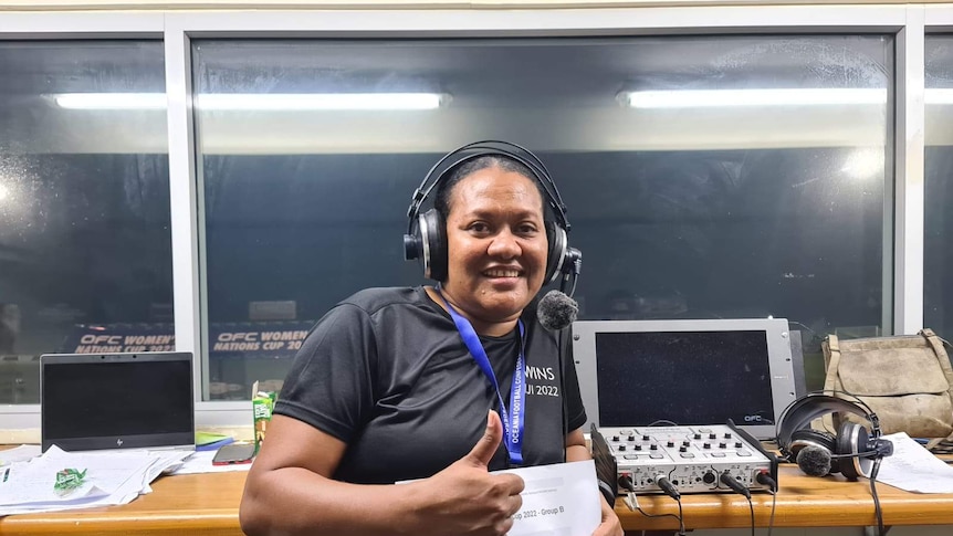 A woman sits at a commentary desk with broadcast gear and wearing headphones, smiling making a thumbs up gesture.