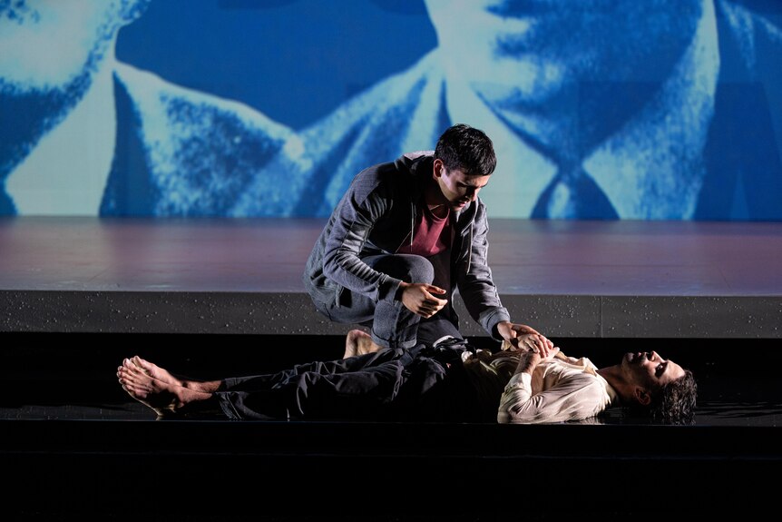 A young man in streetwear crouches over another young man who is lying unconscious onstage.