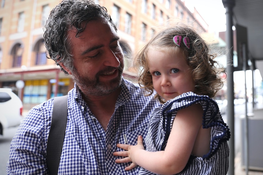 A man in a blue and white shirt holding a toddler in a blue and white dress with watermelon clips in her hair.