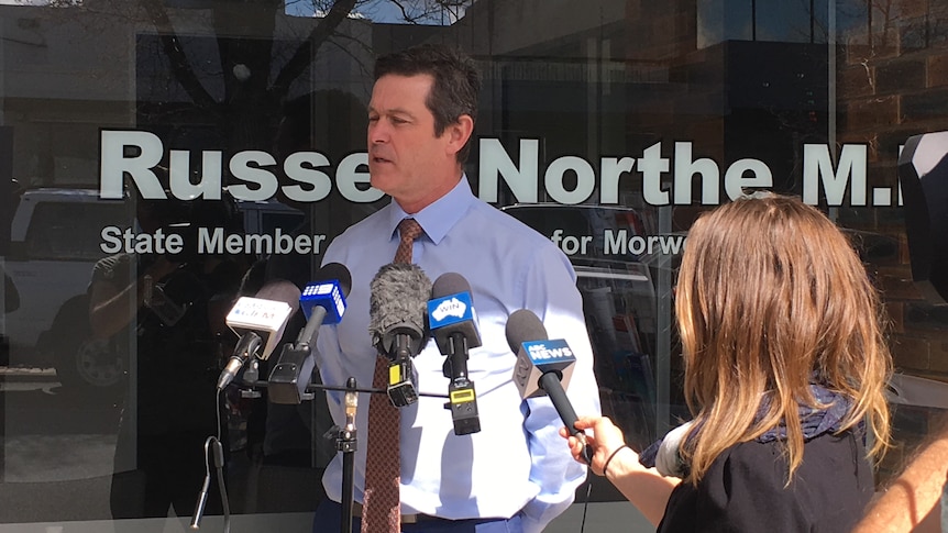 A middle-aged man in a shirt and tie standing behind some microphones at a press conference