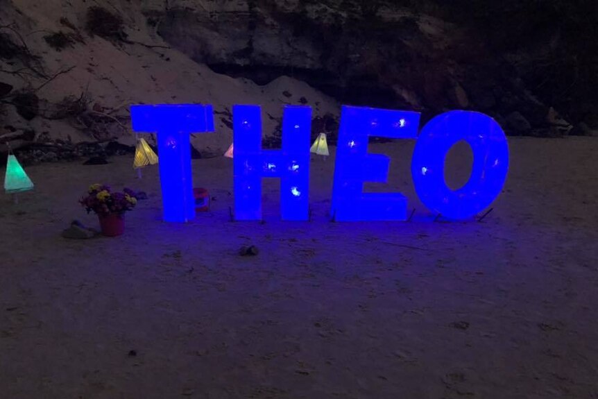 Blue neon blocks spelling 'Theo' in sand.