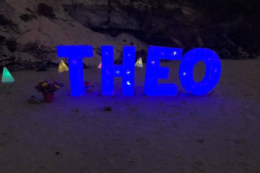 Blue neon blocks spelling 'Theo' in sand.