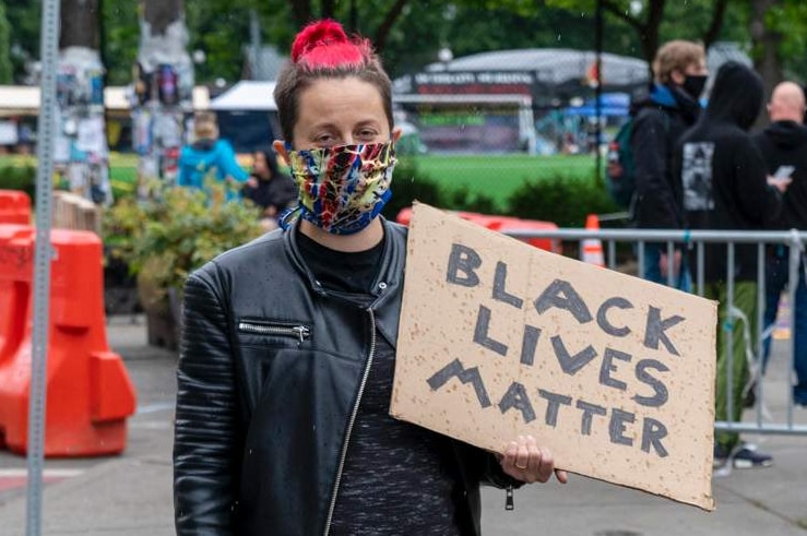 Woman wearing mask, black leather jacket and black to, pink and blonde hair and brown dog holds Black Lives Matter sign