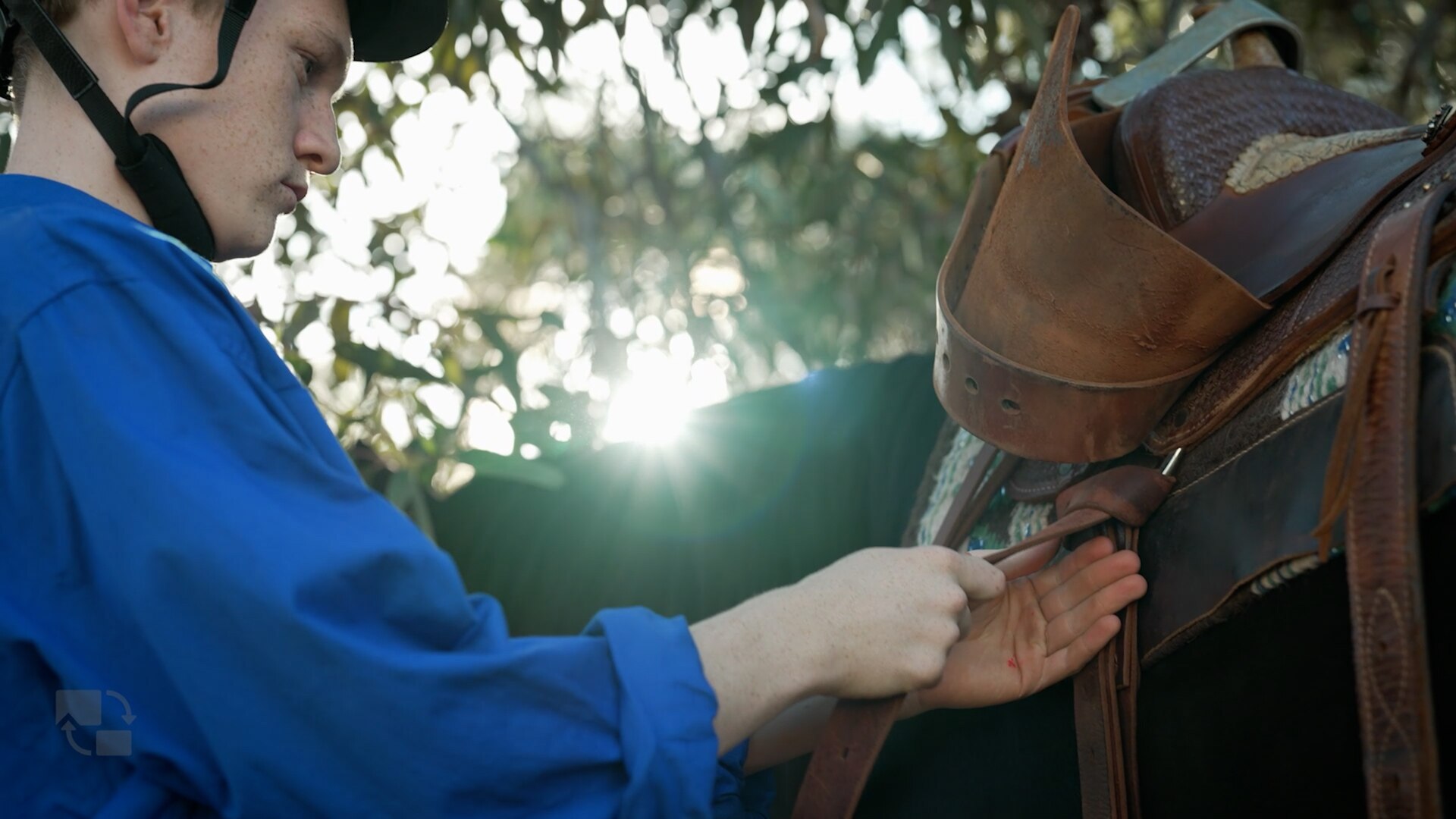 A teenage boy in a long sleeve blue shirt tightening the saddle next to a horse.