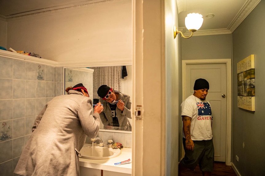 One an stands in a bathroom in front of a mirror brushing his teeth, while another walks through a hallway.