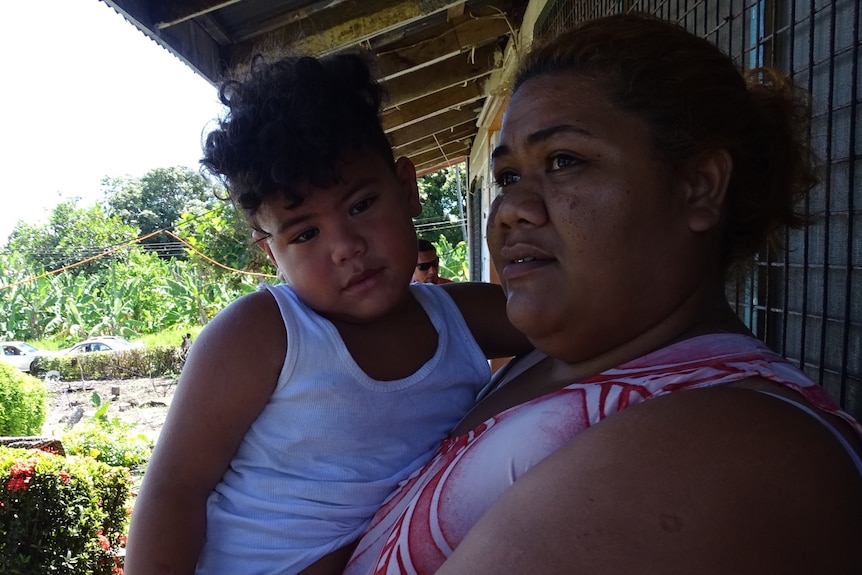 A woman with curly hair holds her son in a singlet and stares off camera outside a building in Samoa.