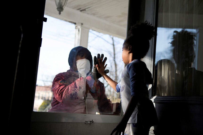 A man in a facemask and gloves puts his hands to a glass door. On the inside, a girl puts her hand to the glass too.