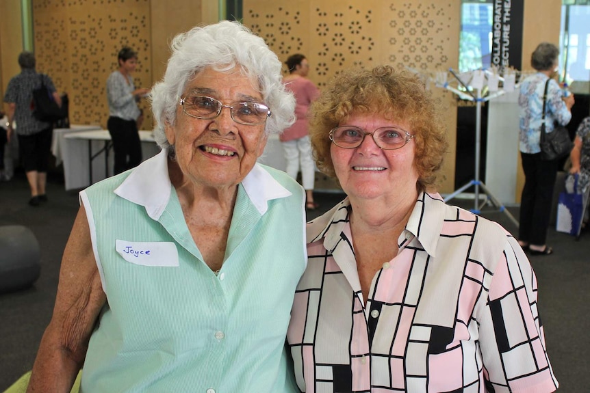 Two older ladies hold each other around the waist while a small conference crowd is in the background