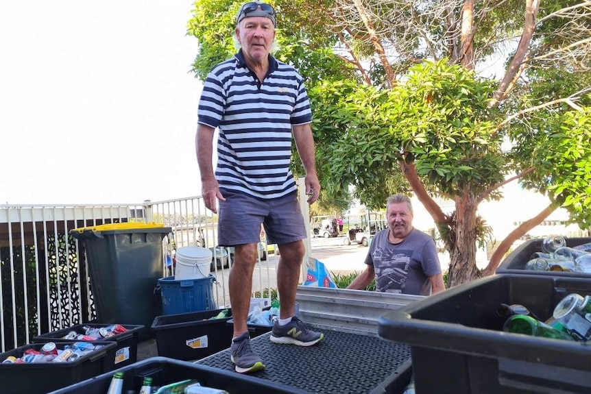 A older man standing on a ute tray surrounded by boxes of empty cans and bottles, with golf buggies in the background