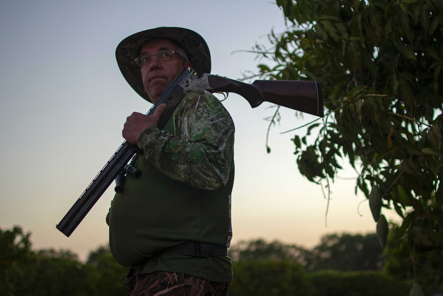 a man in a hat with a gun over his shoulder in a mango orchard.