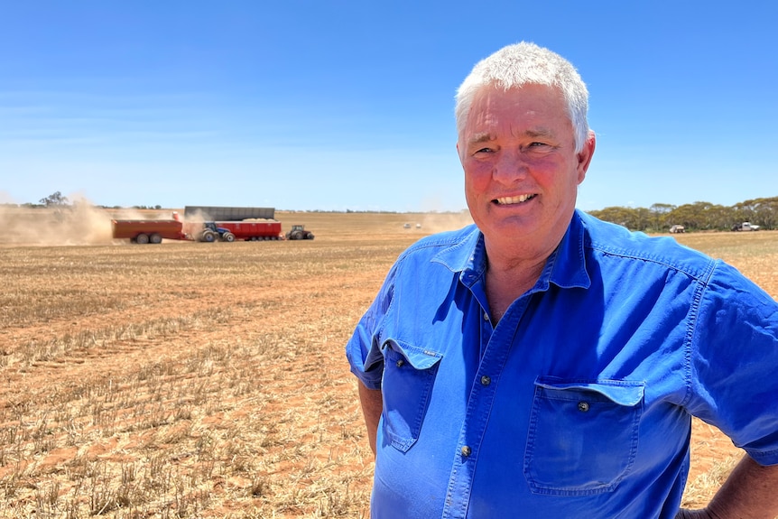 Craig Henderson stands in a paddock and grain machinery can be seen in the background