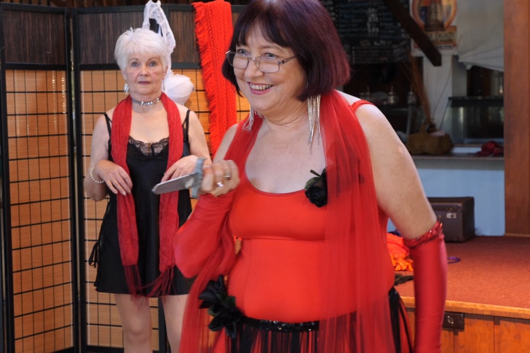 A woman in red holds a pretend dagger pointing it toward the audience with a wicked smile. 