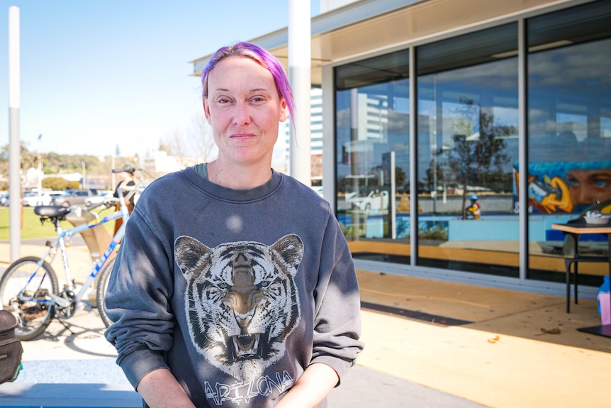 A slightly smiling woman with pink hair sits outdoors in front of a large glass window, wears a jumper with a tiger on it.