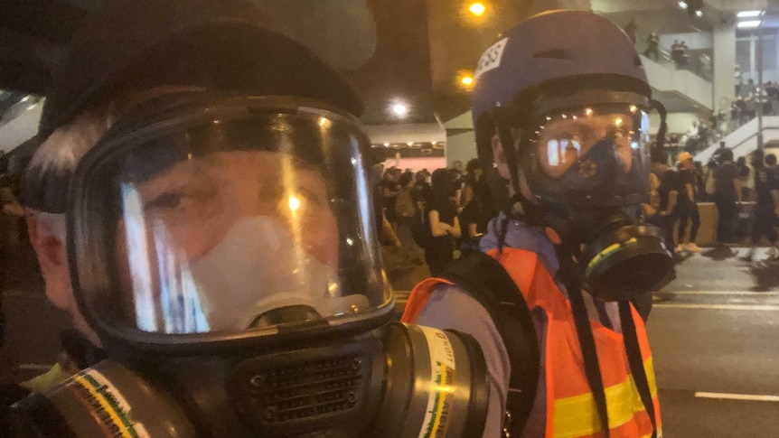 Cameraman and journalist wearing helmets and gasmasks with protestors in background on streets.