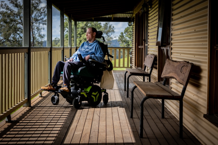 Nathan Johnston sitting in his wheelchair on a large verandah, looking out over a landscape of green hills and leafy trees.