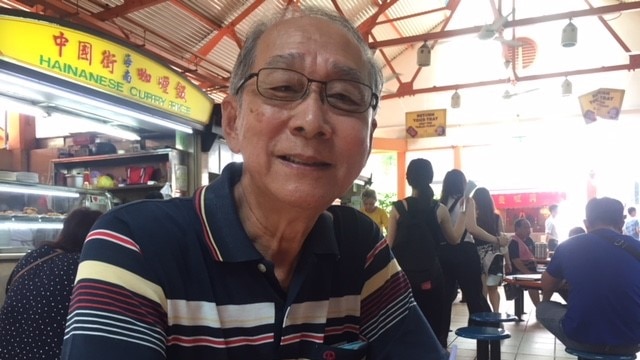 An older man with grey hair and glasses sits in a covered food-court looking at the camera with a half-smile