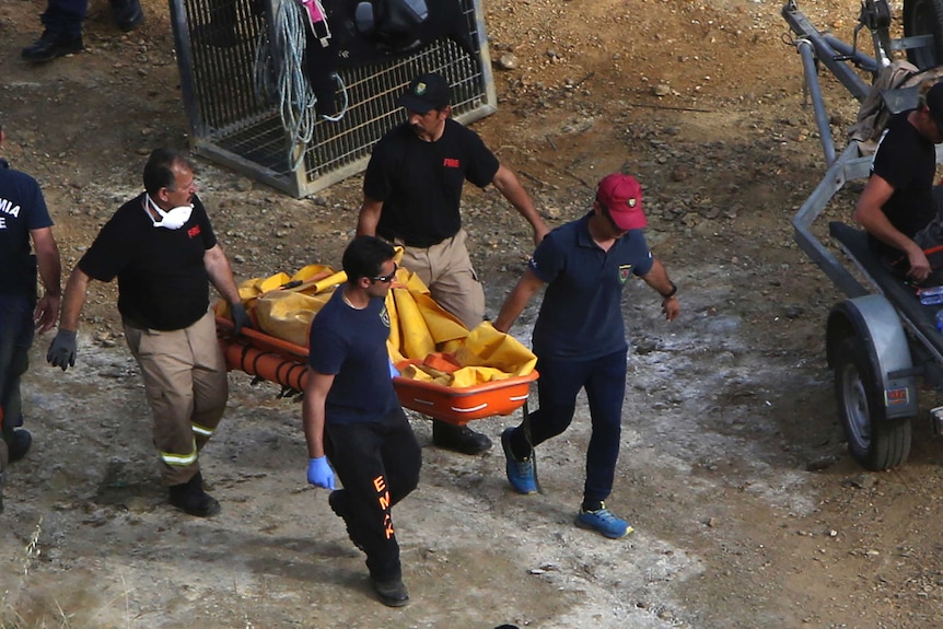 Four men shot from above carry a large orange container with a covered suitcase in it up the bank of a lake.