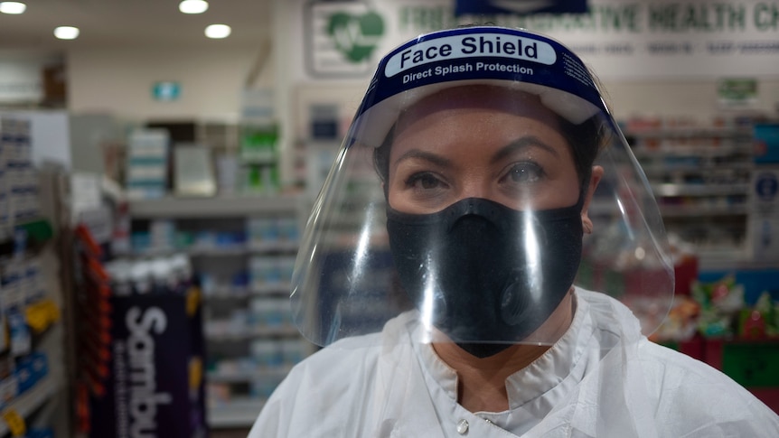 Pharmacist Catherine Bronger wears a white coat and a face shield while inside a pharmacy.