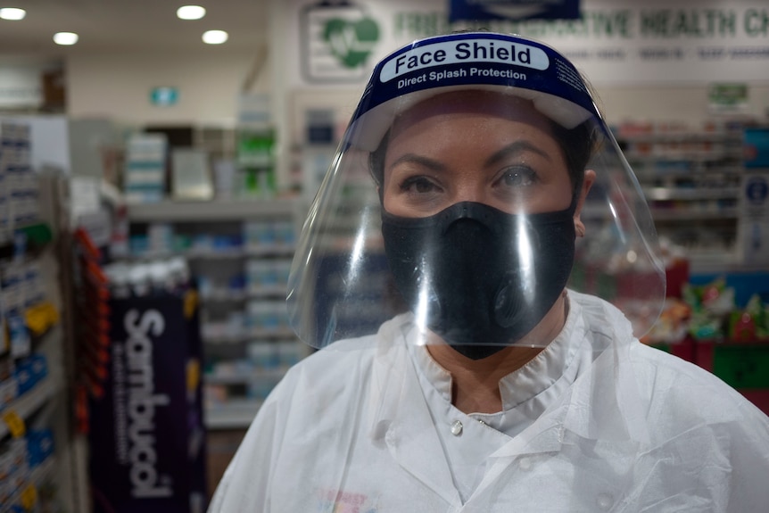 Pharmacist Catherine Bronger wears a white coat and a face shield while inside a pharmacy.