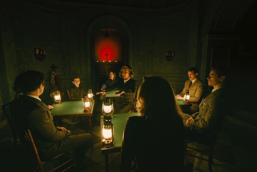 A film still showing a standing woman surrounded by a group of seated people in a dimly lit room with lanterns on tables