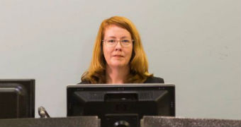 A lady with red hair and glasses looks out from behind a desk and computer.