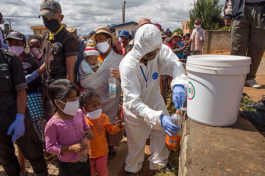 A person in a suit fills a fanta bottle with a brown drink for waiting children in masks.