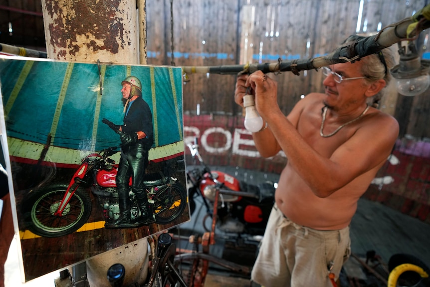Un hombre juguetea con un cable frente a una foto de un hombre en una motocicleta.