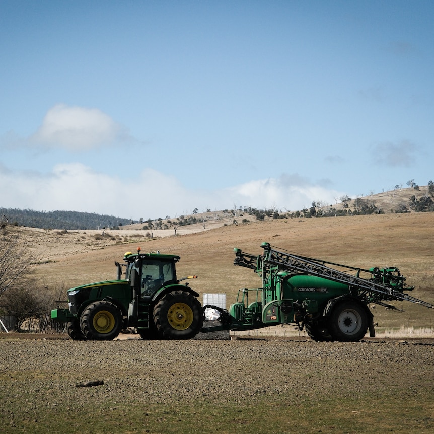 A green tractor towing machienry sits on a very brown-looking farm.