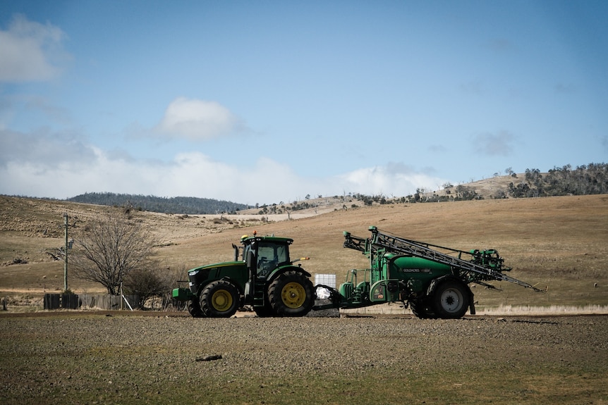 A green tractor towing machienry sits on a very brown-looking farm.