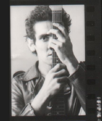 Black and white grainy photo of a young Paul Kelly with spiky dark hair holding the neck of a guitar in front of his face.